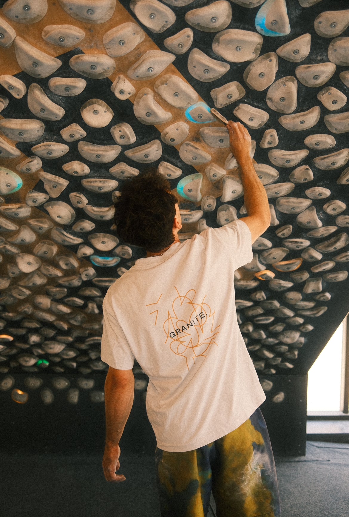 Model wearing white GRANITE. TEE.01 with orange movement lines in the back brushing the kilter board holds in a climbing gym in Copenhagen